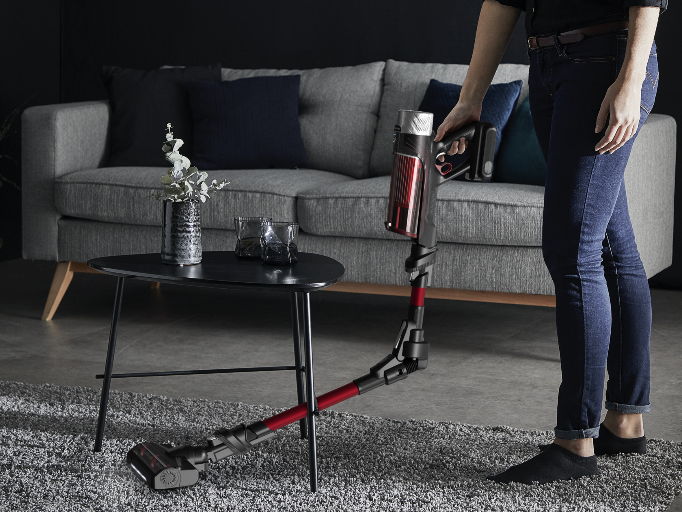 Kobieta odkurza dywan czerwono-czarnym odkurzaczem. Szara sofa i czarny stolik w tle.