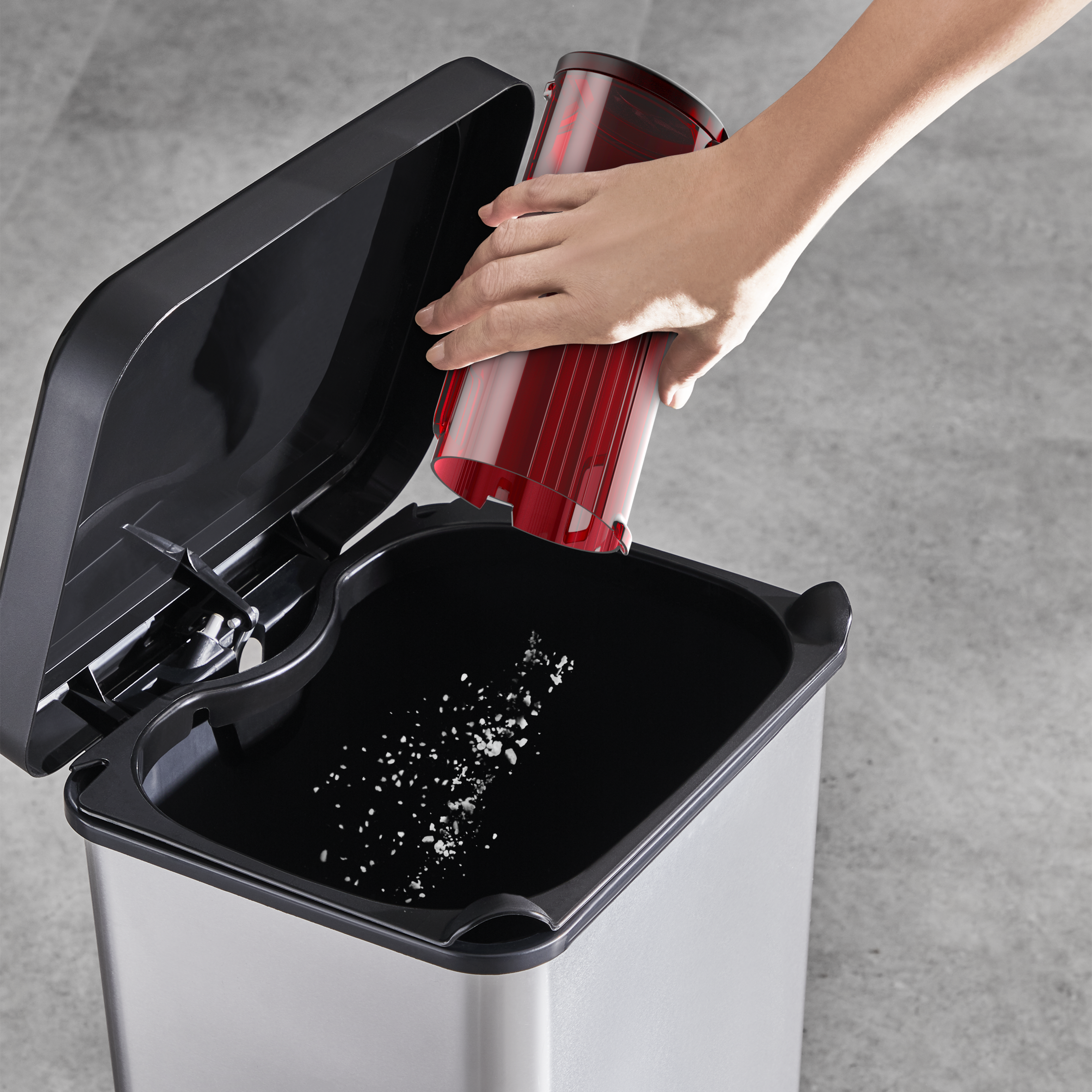 Ręka opróżniająca czerwony pojemnik na kurz do srebrnego kosza na śmieci. Szara podłoga w tle.