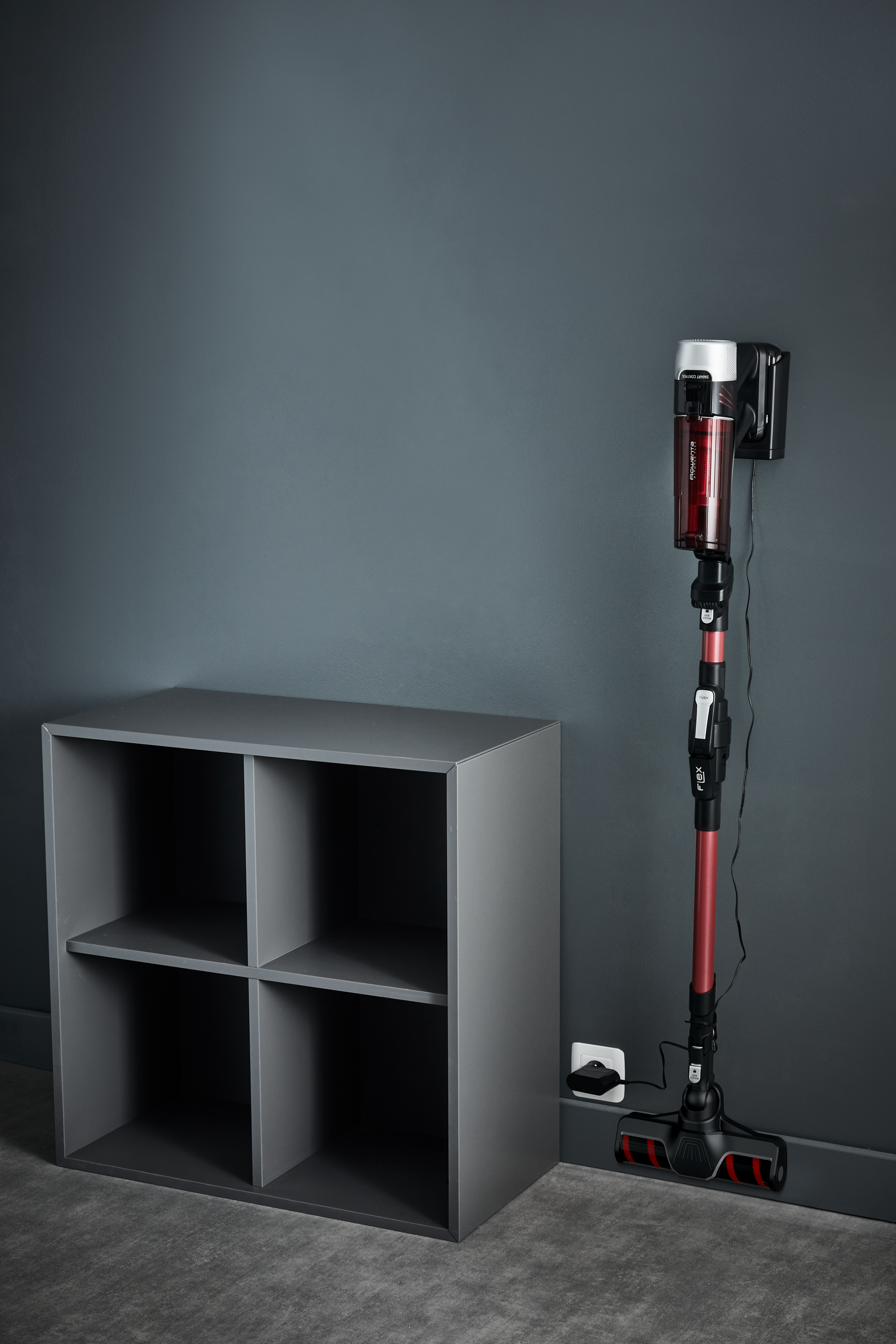 Bezprzewodowy odkurzacz w trakcie ładowania, umieszczony obok szarej półki na szarej ścianie. Widoczne gniazdo zasilania.