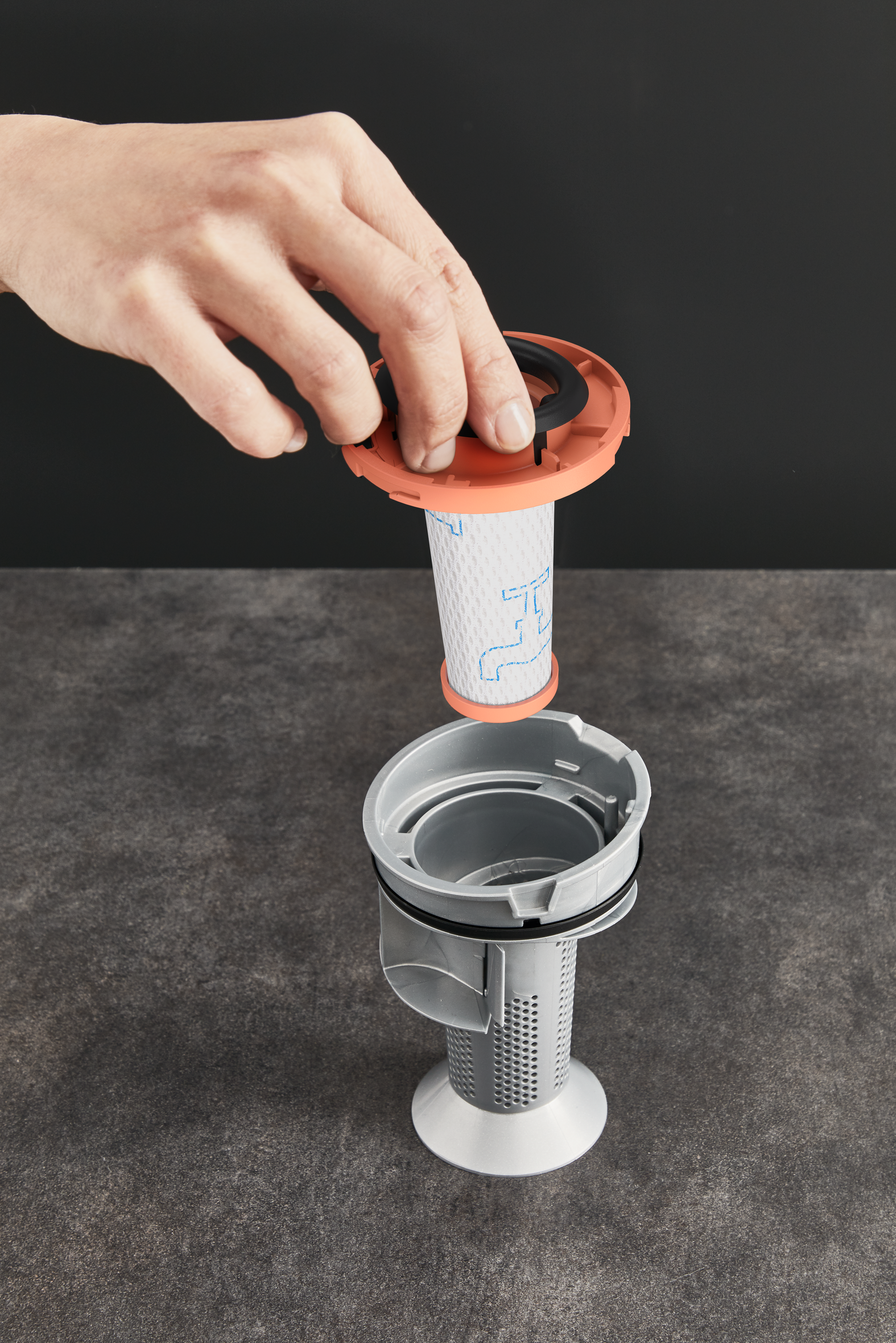 Ręka trzymająca pomarańczową nasadkę filtra nad biało-szarym zespołem filtracyjnym. Ciemnoszara powierzchnia.
