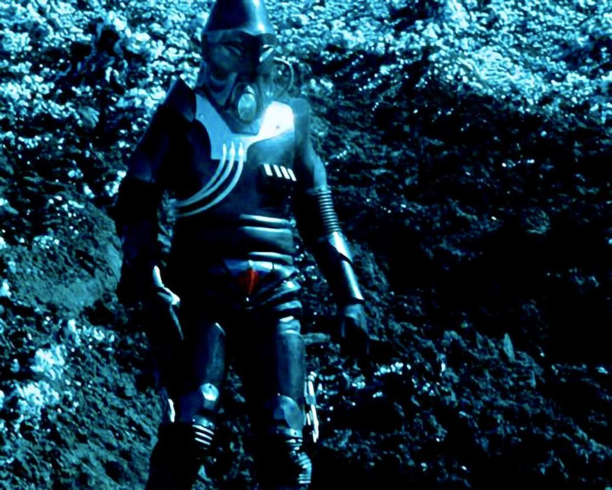 Eine Figur in einem schwarzen Anzug und Helm mit Atemschutzgerät steht auf einer felsigen, blau getönten Oberfläche.