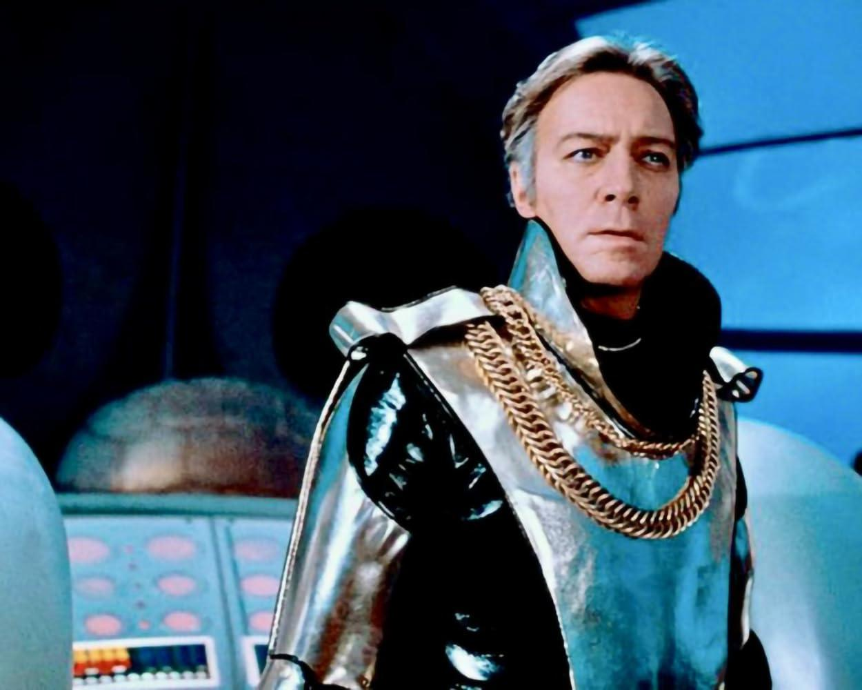 Ein Mann in einem silbernen Outfit mit einer Goldkette steht in einem futuristischen Raum und wirkt besorgt.