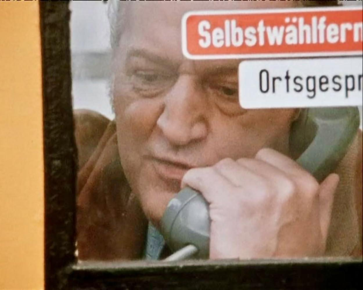 Mann in Telefonzelle. Er wirkt besorgt und hält ein graues Telefon ans Ohr. Rotes Schild: Selbstwahl.