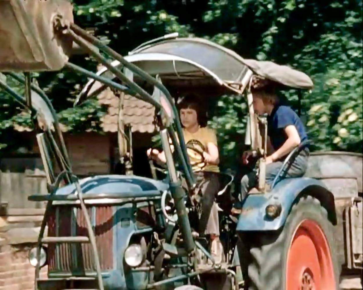 Zwei Jungen auf einem blauen Traktor auf einem Feld. Einer fährt, der andere sitzt daneben. Bäume im Hintergrund.