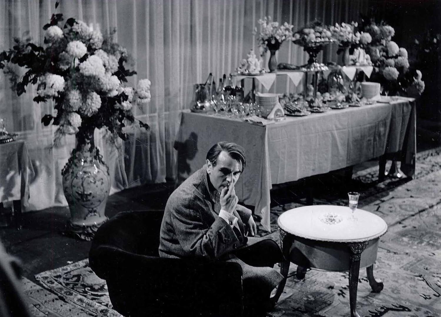 Mann im Anzug, sitzt, Hand am Kinn. Tisch mit Essen, Blumen. Hintergrund: andere Leute, verzierte Einrichtung.