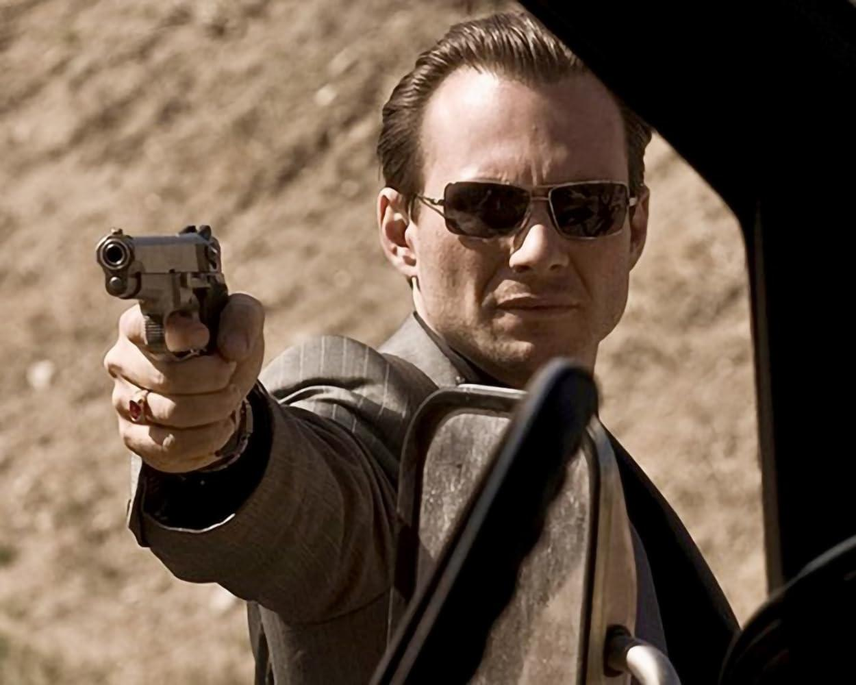 Mann mit Sonnenbrille zielt mit Waffe. Er sitzt in einem Auto. Wüstenlandschaft im Hintergrund.