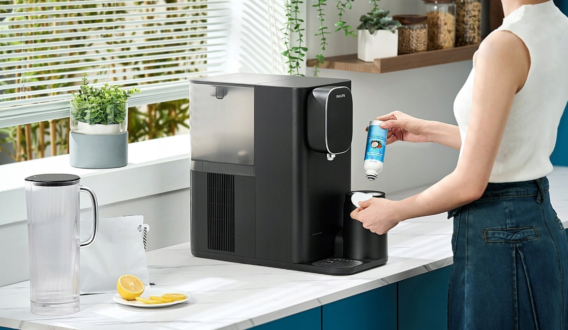 Osoba dodaje filtr do czarnego dystrybutora wody. Szklanka i cytryna na blacie są w tle.