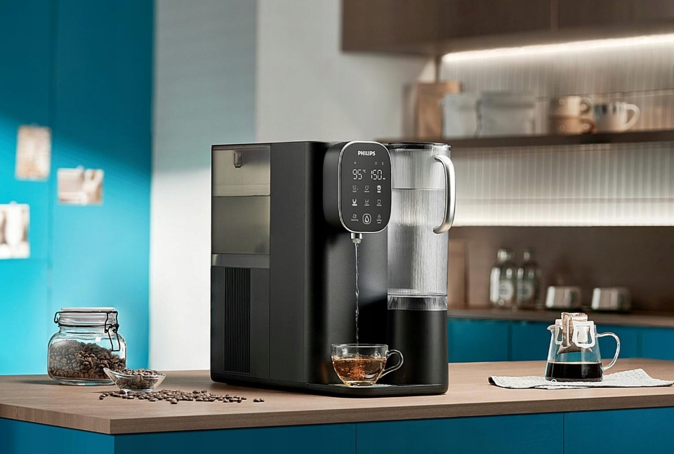 Czarny dystrybutor wody Philips z filiżanką herbaty. Ziarna kawy i ekspres do kawy są w tle.