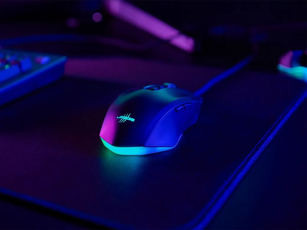 Fekete gamer egér zöld díszítéssel, sötét felületen, kék és lila fénnyel megvilágítva. Billentyűzet részben látható.