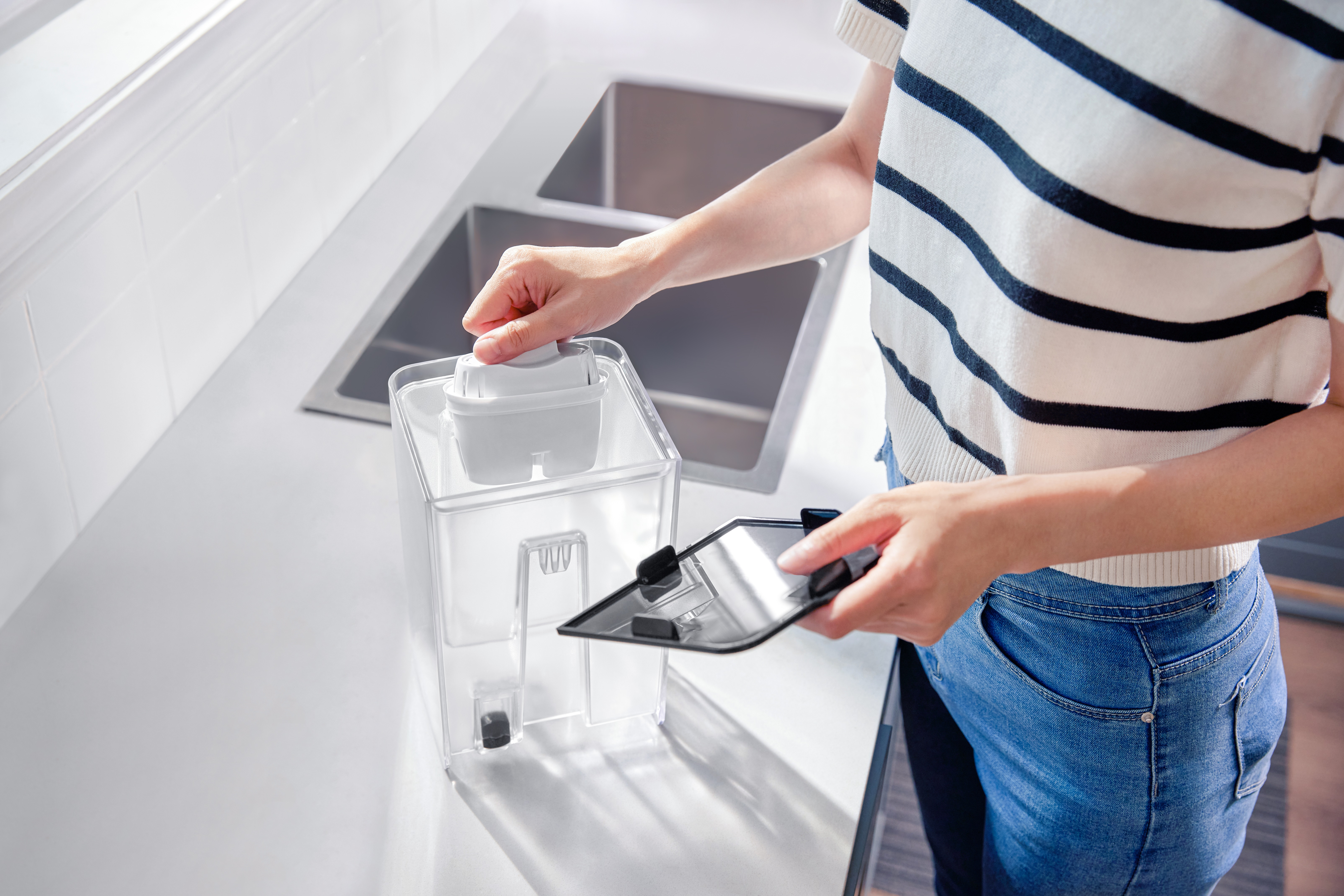 Osoba wyjmuje filtr wody z dozownika. Biały blat i zlew w tle. Biało-czarny filtr.