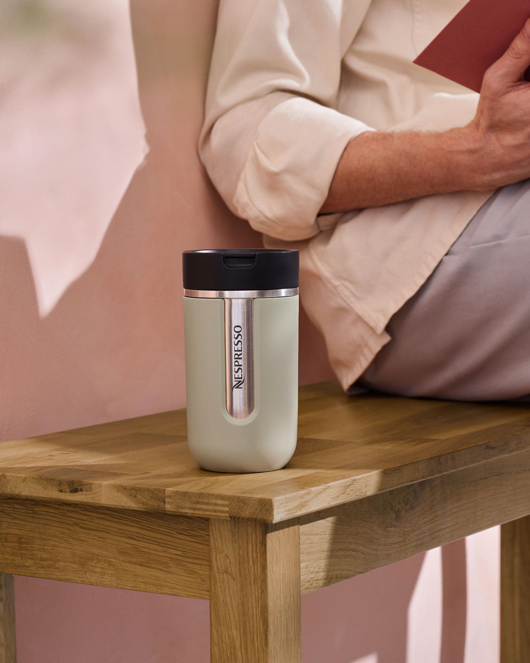 Ein grüner Nespresso-Reisebecher auf einer Holzbank. Im Hintergrund liest eine Person ein Buch.