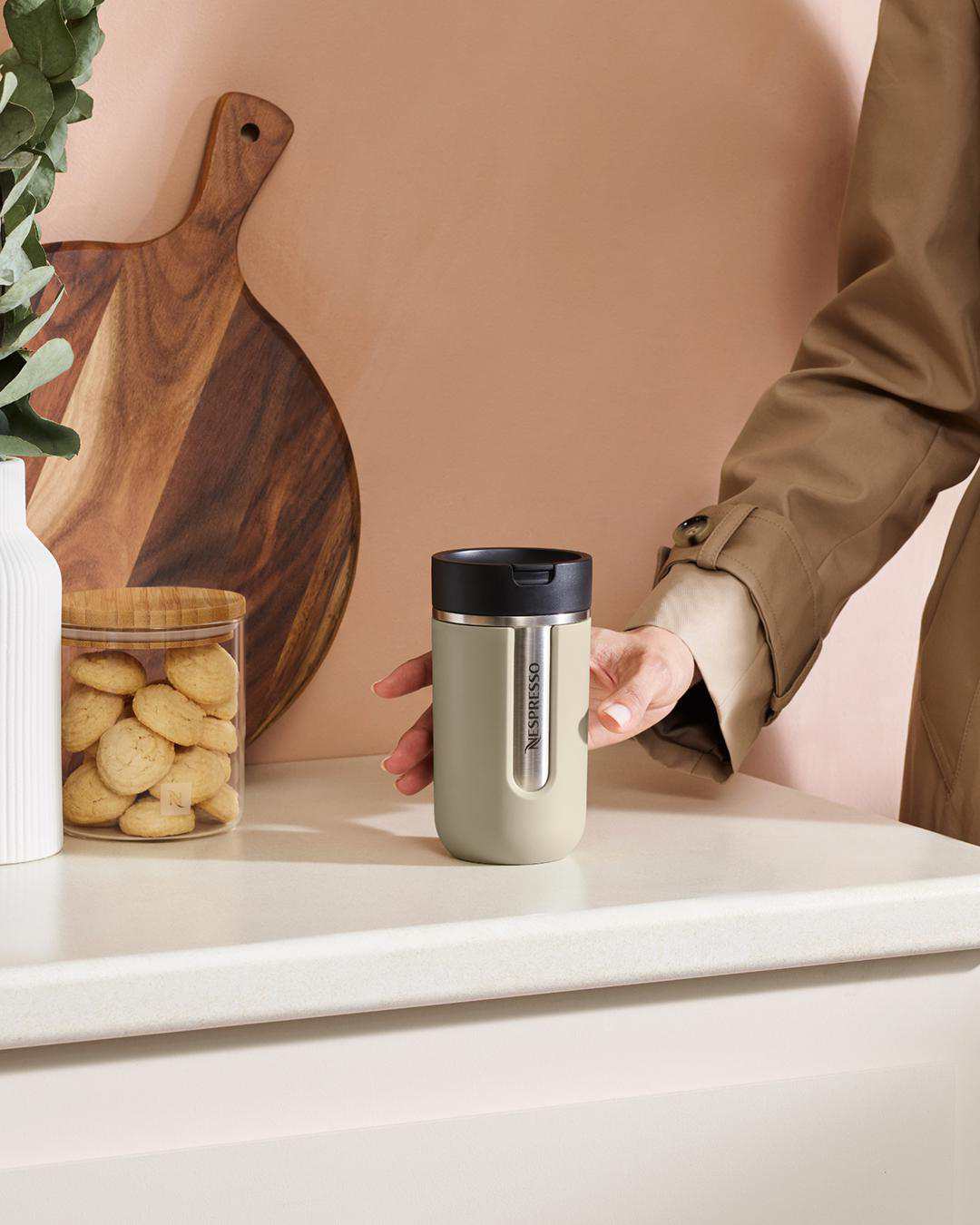 Eine Hand greift nach einem grünen Nespresso-Reisebecher auf einer weißen Arbeitsplatte. Ein Schneidebrett ist im Hintergrund.