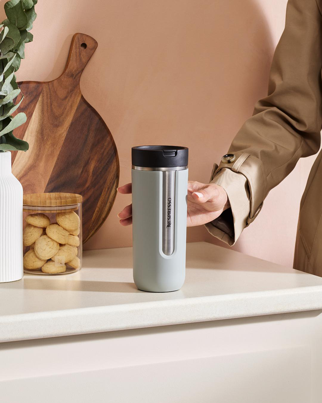 Person hält hellgrauen Nespresso-Reisebecher, steht an weißer Theke mit Deko.