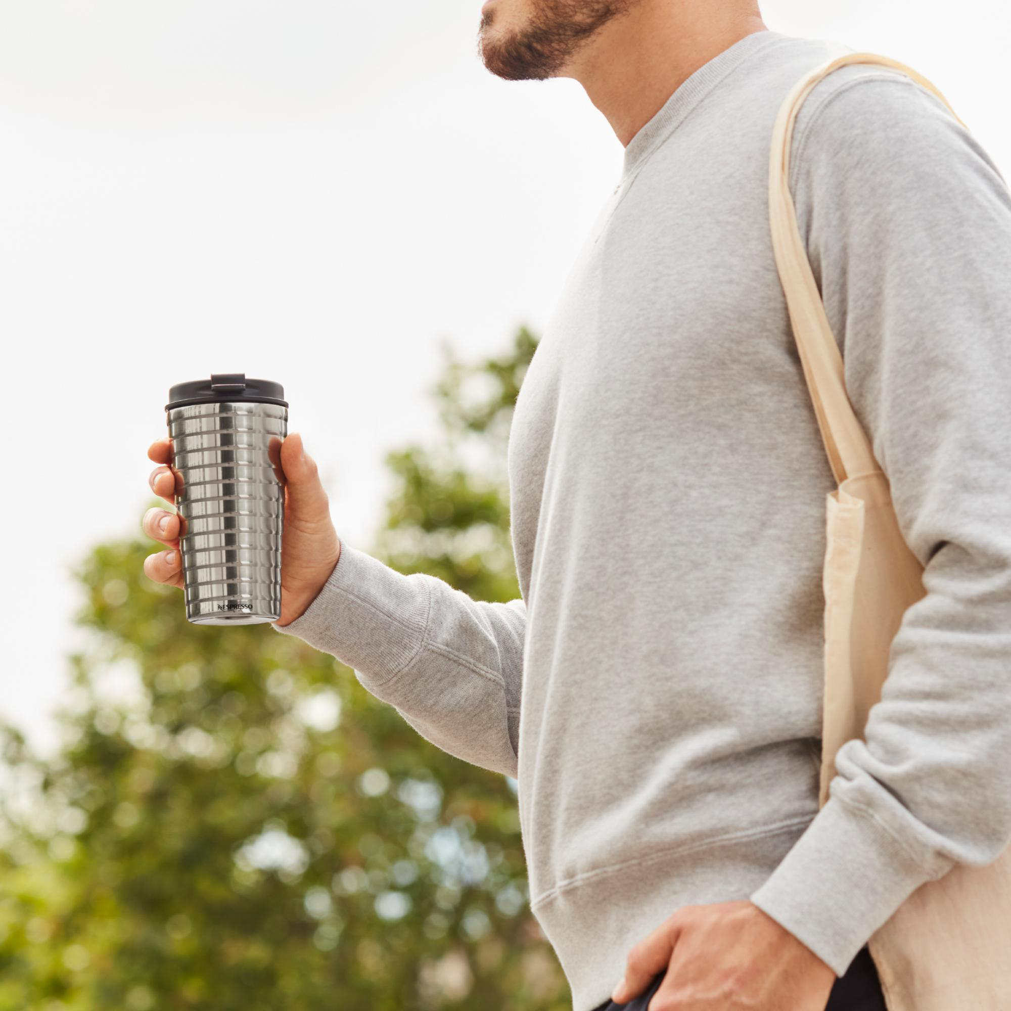 Mann hält Edelstahl-Nespresso-Becher im Freien, trägt Tragetasche. Graues Sweatshirt.