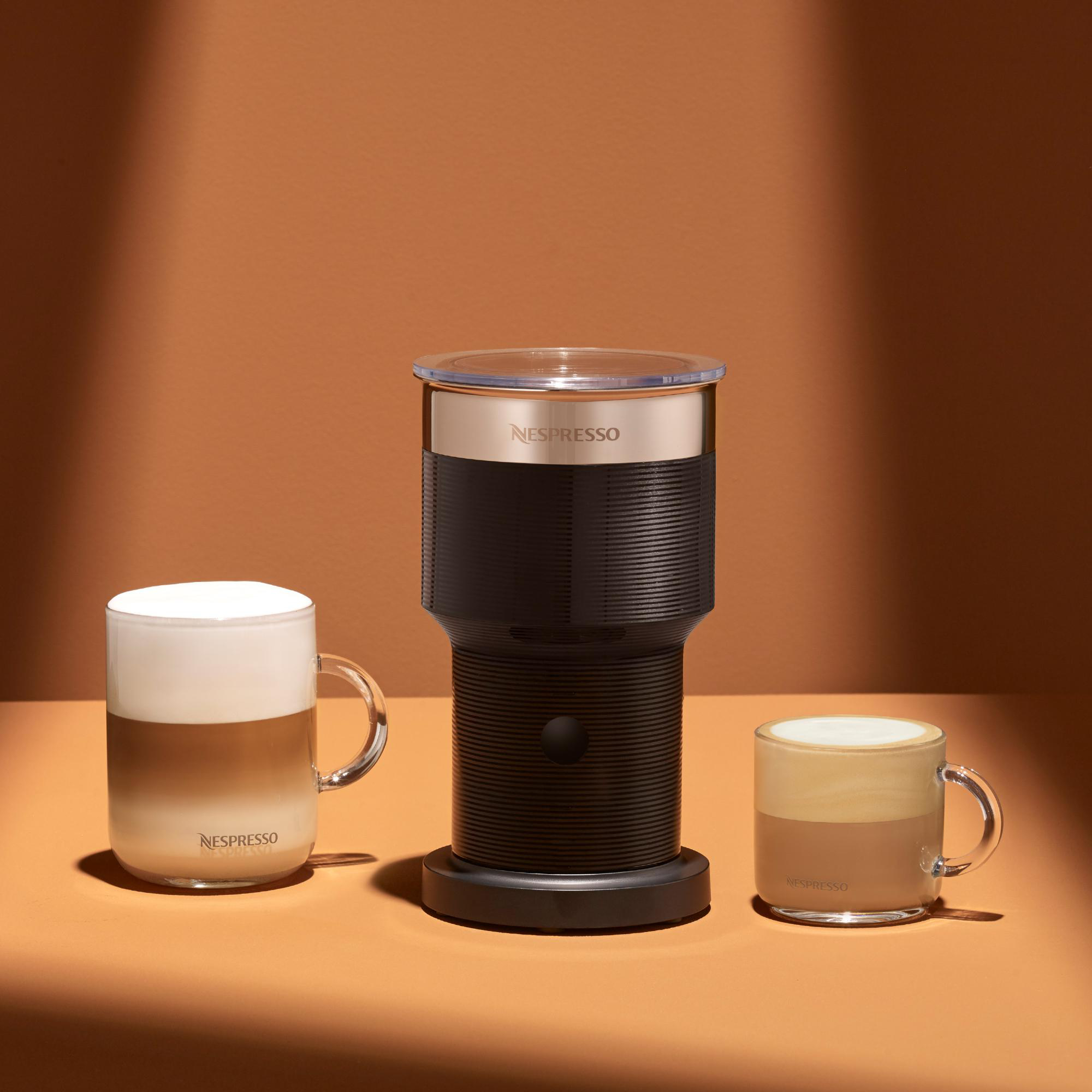 Nespresso-Milchaufschäumer mit zwei Kaffeetassen auf orangefarbenem Hintergrund. Eine Tasse hat geschichteten Kaffee, die andere Schaum.
