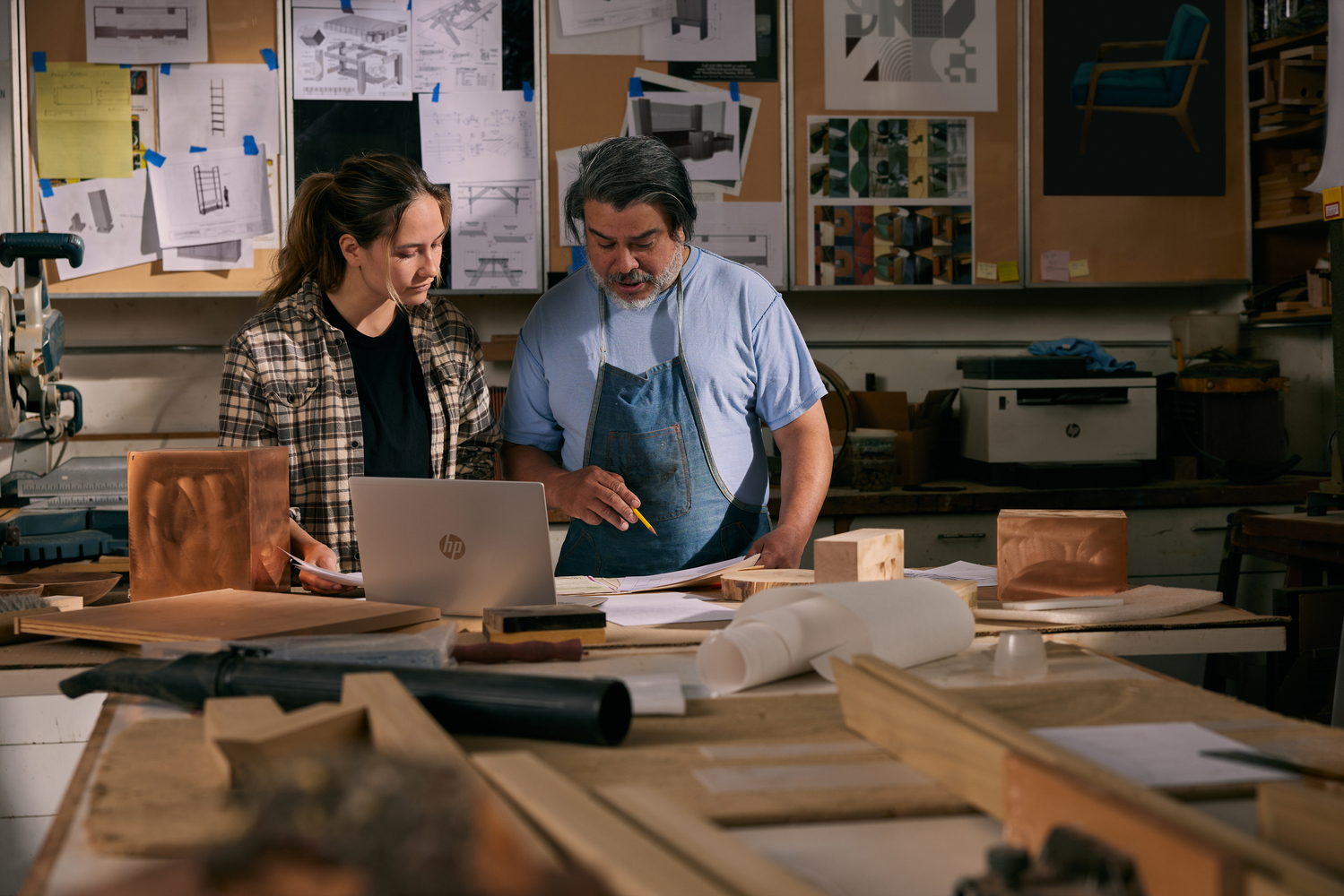 Dos personas mirando un portátil en un taller. Madera y herramientas al fondo.