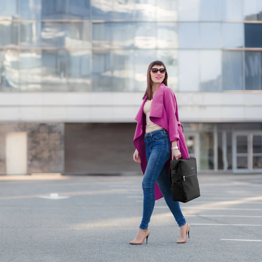 Kobieta w różowym płaszczu idzie ulicą. Czarny plecak w dłoni. Nowoczesny budynek w tle.