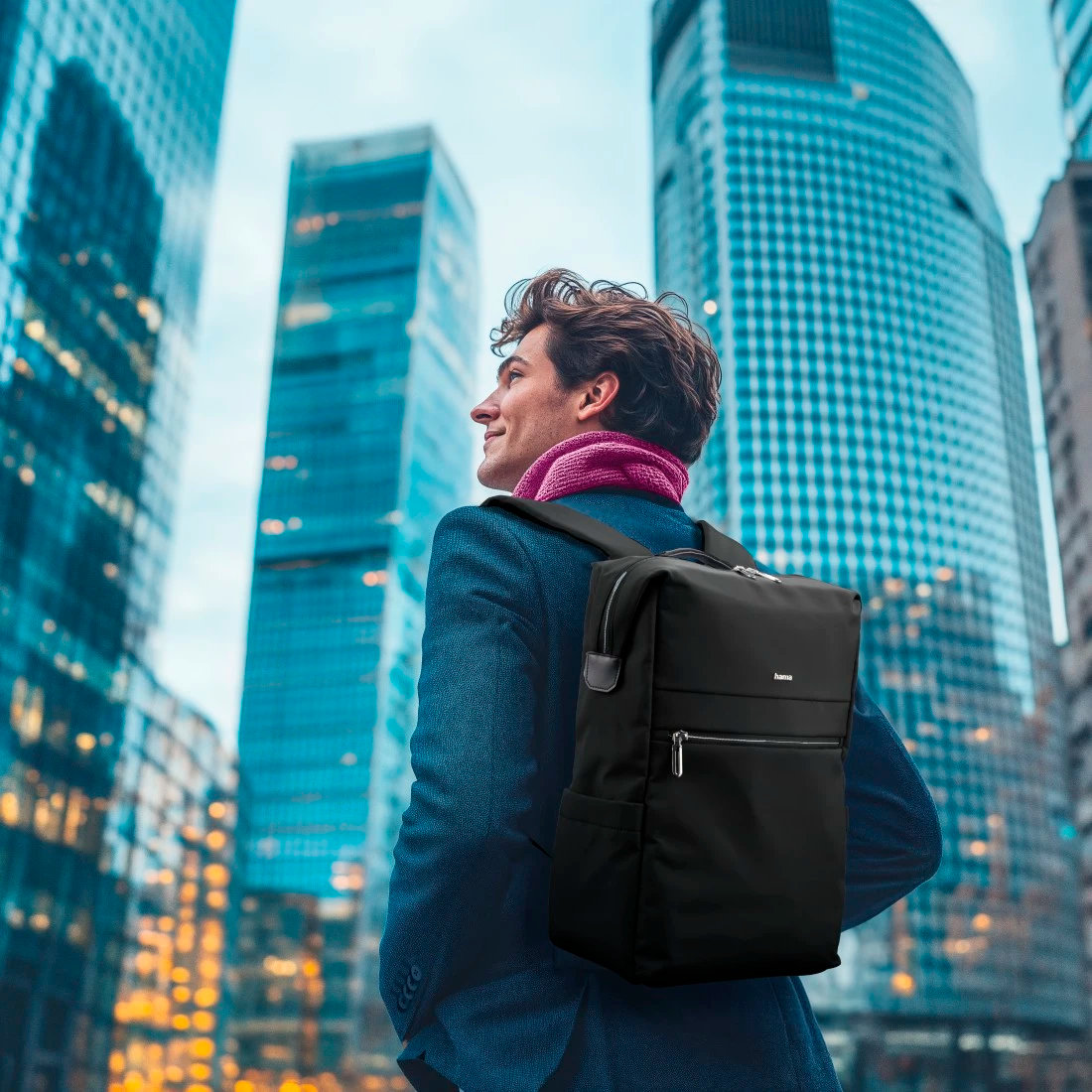 Mężczyzna w niebieskim garniturze z czarnym plecakiem. Na tle drapaczy chmur. Różowy szalik na szyi.