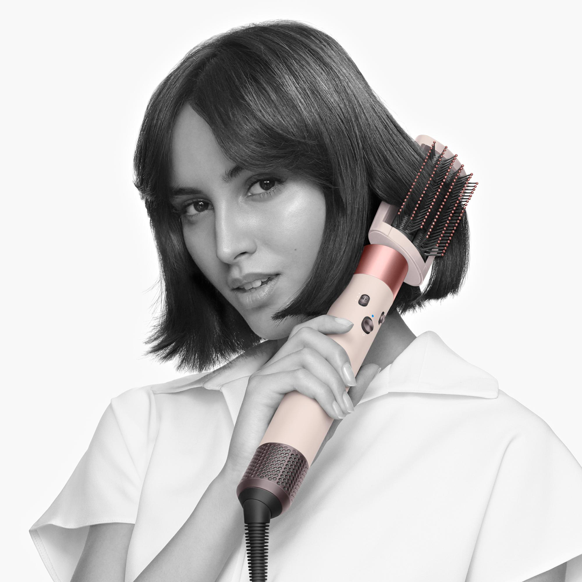 Eine Frau mit kurzen dunklen Haaren hält ein Haarstyling-Gerät. Sie trägt ein weißes Hemd. Der Hintergrund ist weiß.