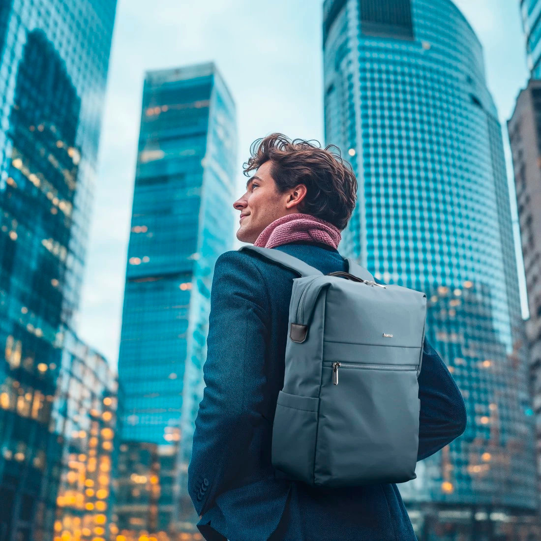 Mężczyzna w mieście, niosący szary plecak, w szaliku i płaszczu, z wysokimi budynkami.