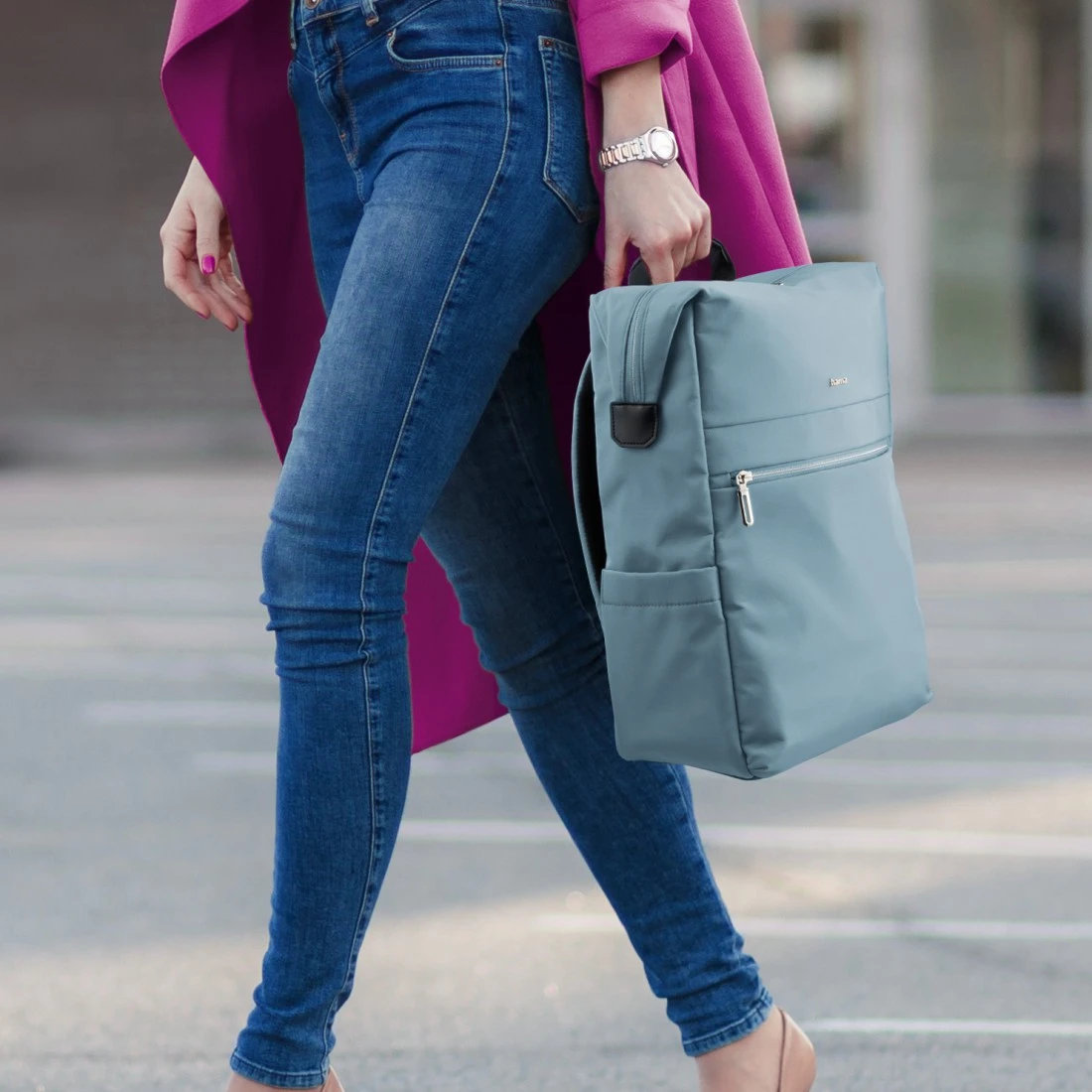 Kobieta idzie, turkusowy plecak, dżinsy, różowy płaszcz i zegarek. Niewyraźne tło.