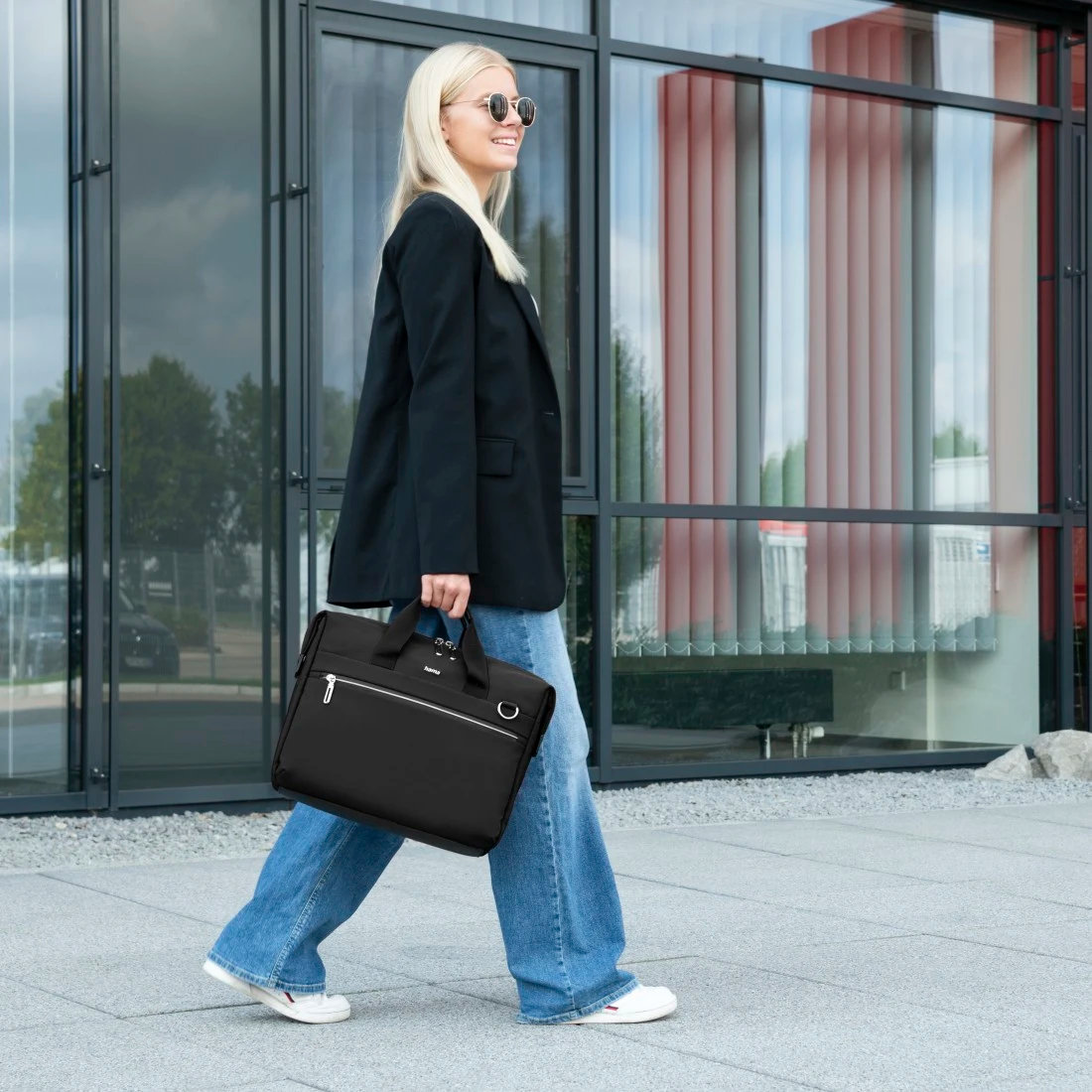 Kobieta idzie z czarną teczką. Ma na sobie marynarkę i dżinsy. Budynki w tle.