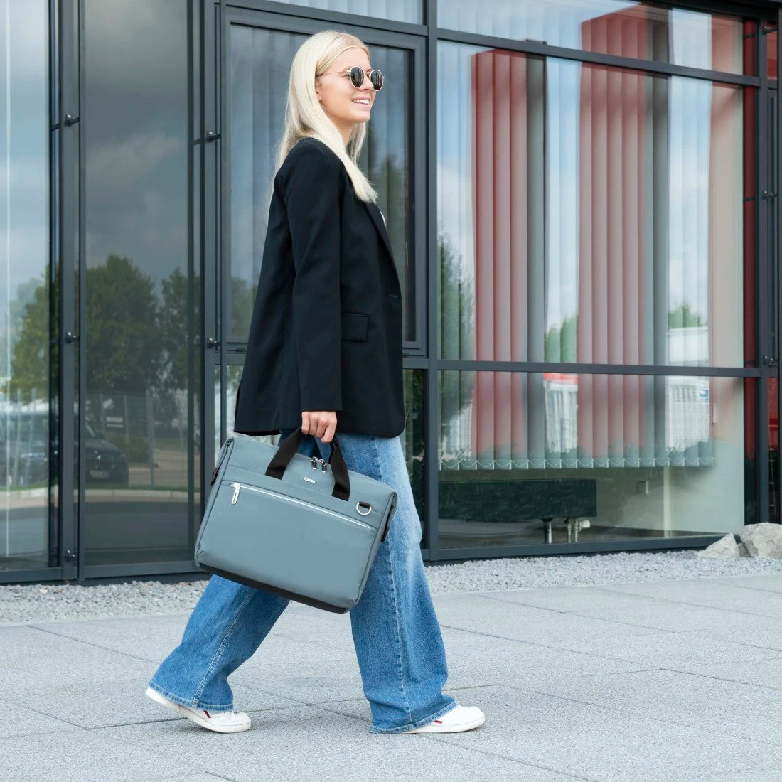 Kobieta idąca, niosąca szarą torbę, w czarnej marynarce, niebieskich dżinsach i okularach przeciwsłonecznych. Szklany budynek.