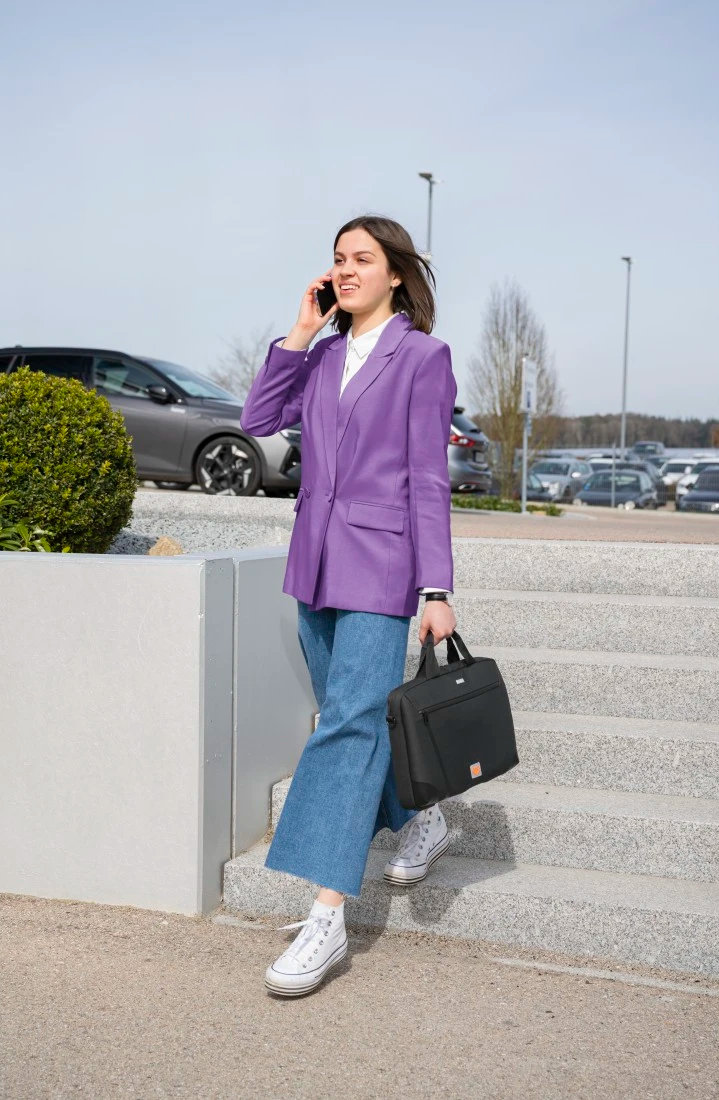 Kobieta w fioletowej marynarce i dżinsach schodzi po schodach, rozmawia przez telefon, trzyma torbę. Samochody i drzewa w tle.