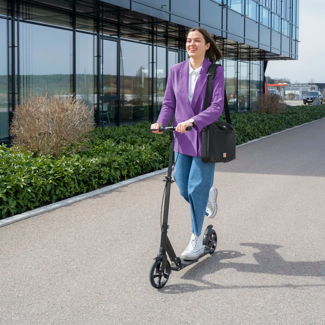 Kobieta w fioletowej marynarce jedzie na czarnej hulajnodze po ścieżce. Ma torbę i uśmiecha się. W tle budynek i zieleń.