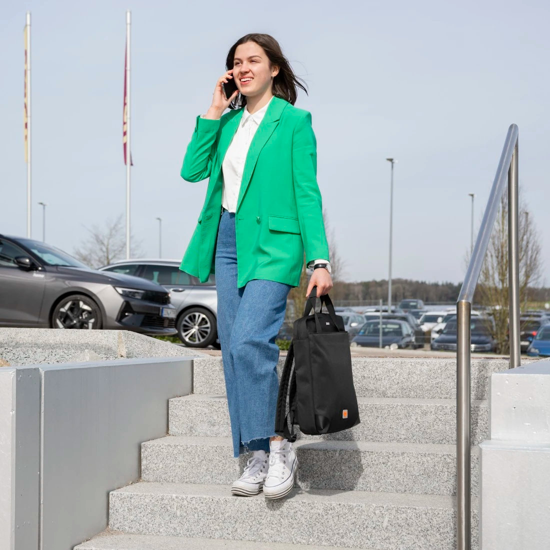 Kobieta w zielonej kurtce i dżinsach, trzymająca czarną torbę, rozmawiająca przez telefon.