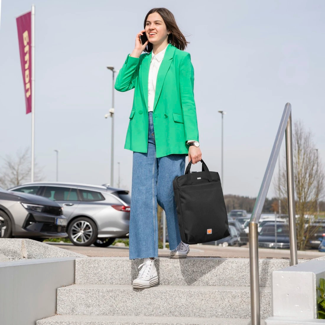 Kobieta w zielonej kurtce i dżinsach, trzymająca czarną torbę, rozmawiająca przez telefon, idąca.