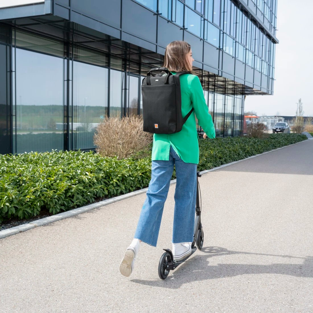 Kobieta w zielonej marynarce, dżinsach, jedzie na czarnym skuterze, czarny plecak, obok budynku.