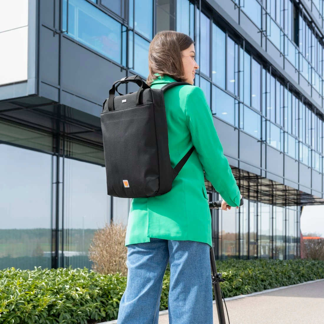 Kobieta w zielonej marynarce, dżinsach, czarny plecak, jedzie na czarnym skuterze, obok budynku.