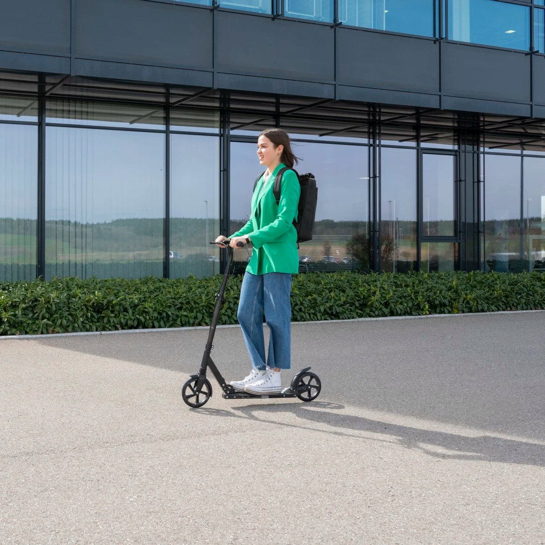Kobieta w zielonej marynarce i dżinsach jedzie czarnym skuterem obok budynku, z czarnym plecakiem.