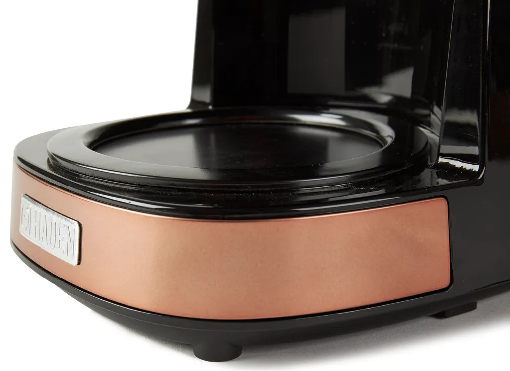 Zbliżenie podstawy ekspresu do kawy. Miedziane wykończenie, czarny korpus. Widoczna nazwa marki.