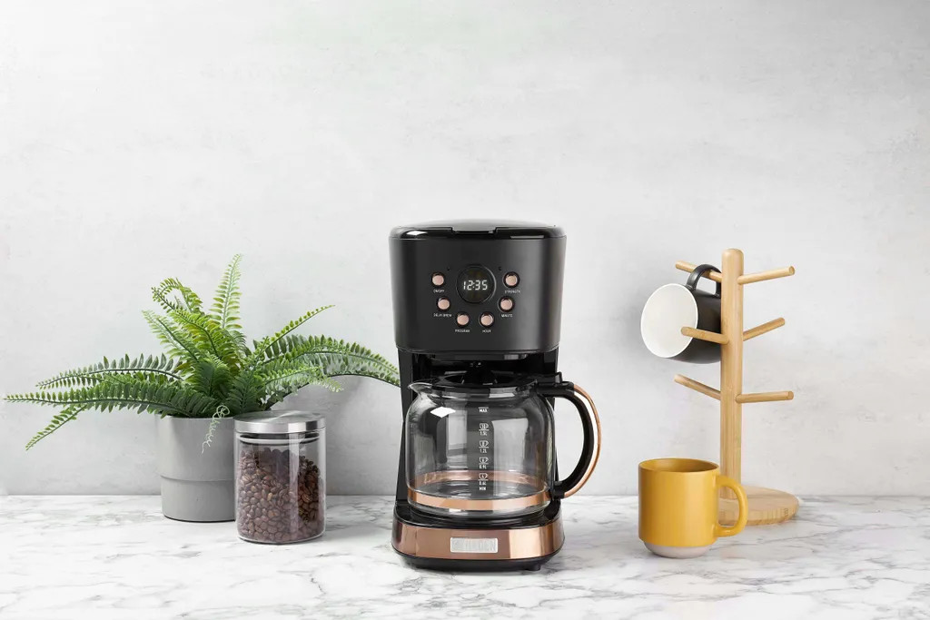 Ekspres do kawy na blacie z ziarnami kawy, rośliną, kubkiem i drewnianym uchwytem na kubki w kształcie drzewa.