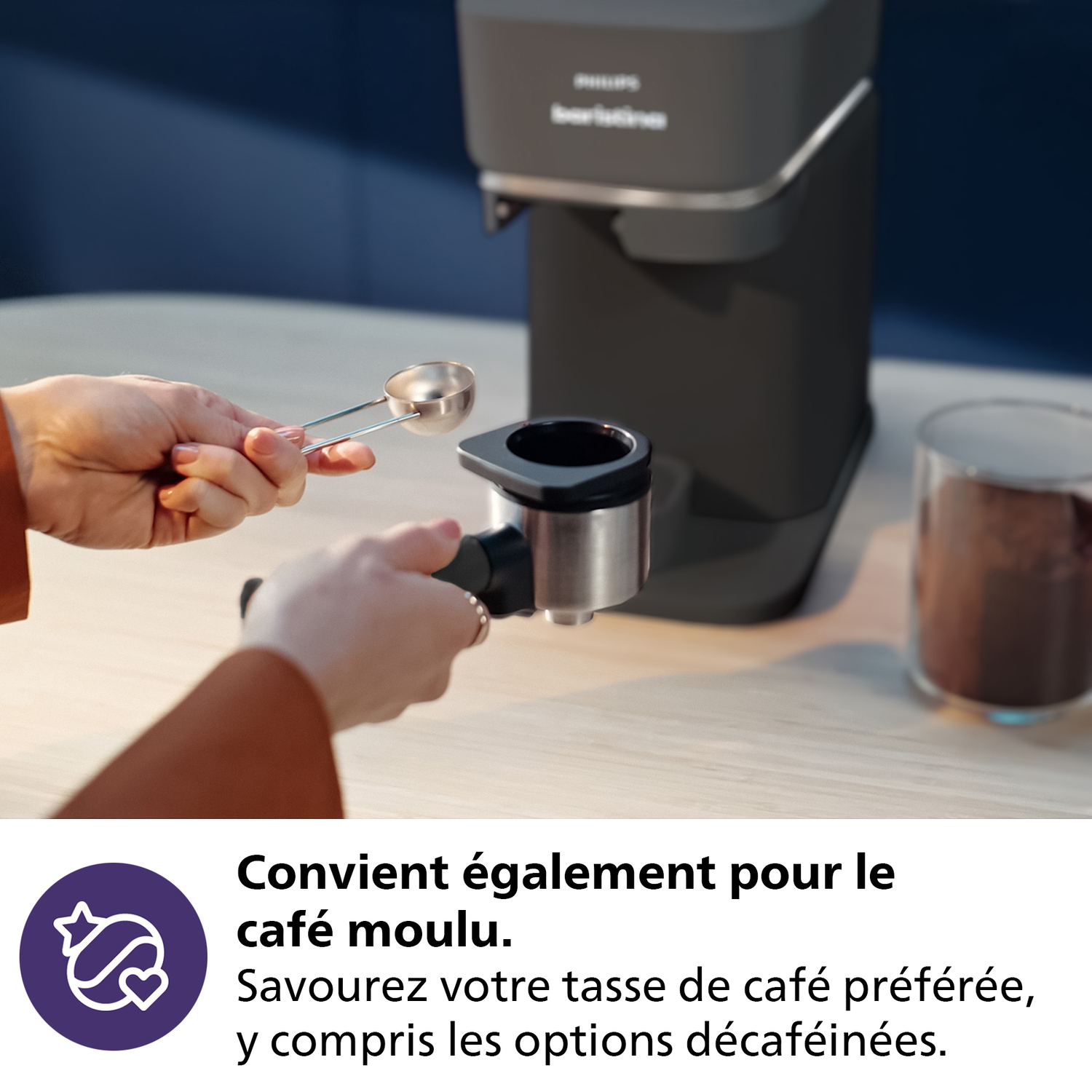 Main avec cuillère ajoute du café au porte-filtre. Machine à café noire, pot en verre. Comptoir bois.