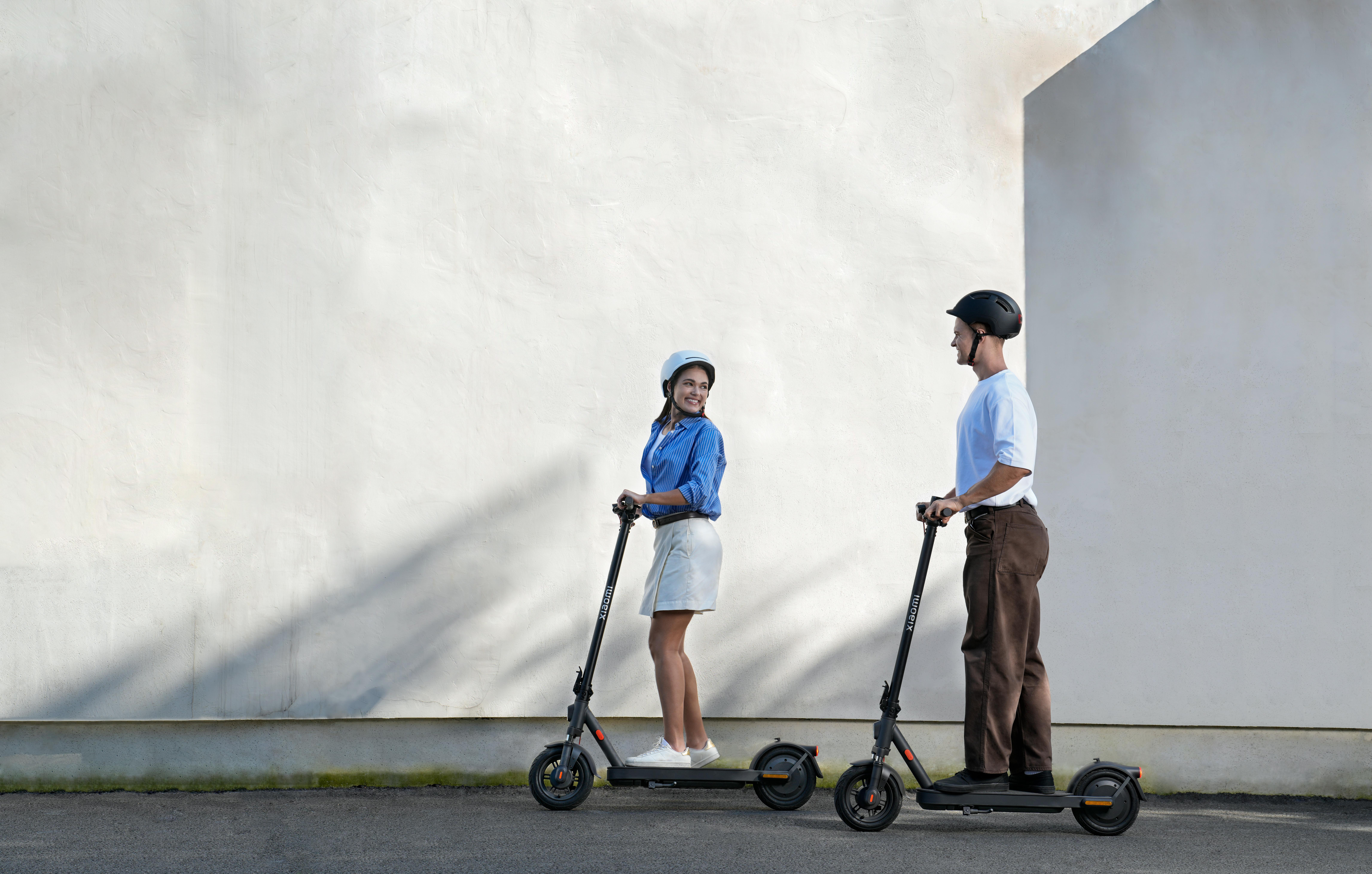 Zwei Personen fahren auf schwarzen Xiaomi-Rollern vor einer weißen Wand. Sie tragen Helme.