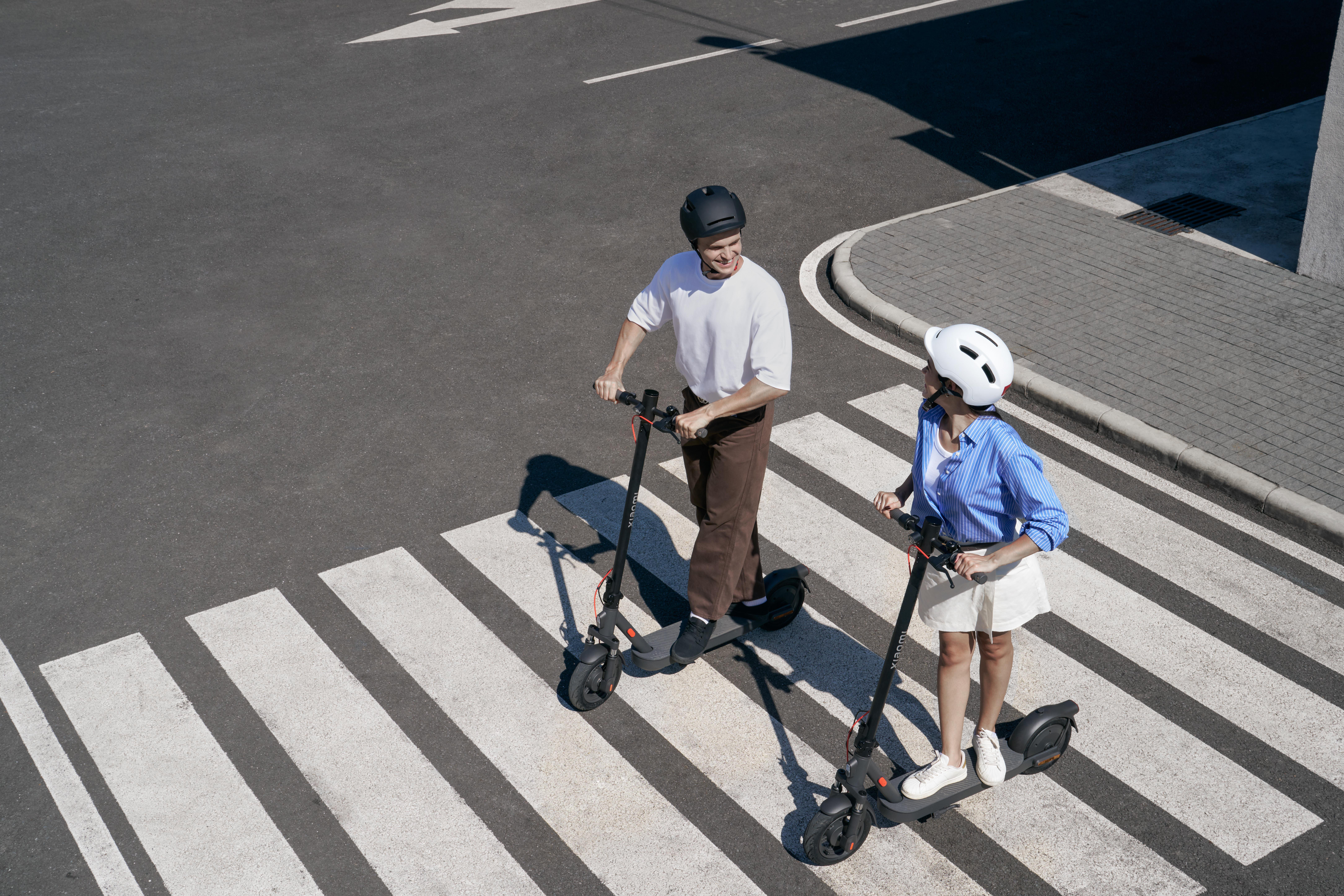 Zwei Personen fahren mit Elektrorollern auf einem Zebrastreifen. Sie tragen Helme.