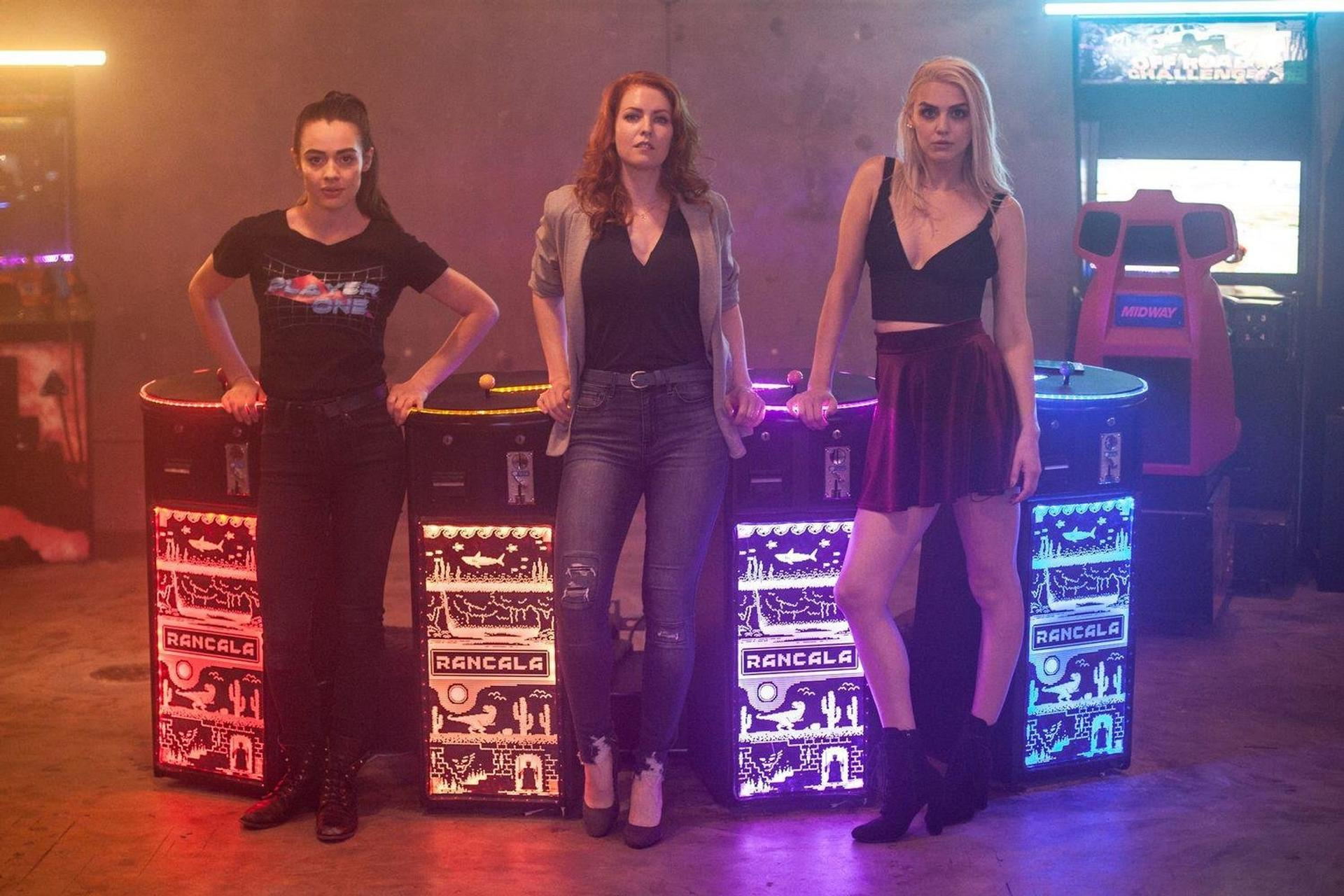 Drei Frauen posieren an Spielautomaten. Eine trägt ein Player One-Shirt. Die Automaten tragen "Rancala". Neonlichter im Hintergrund.