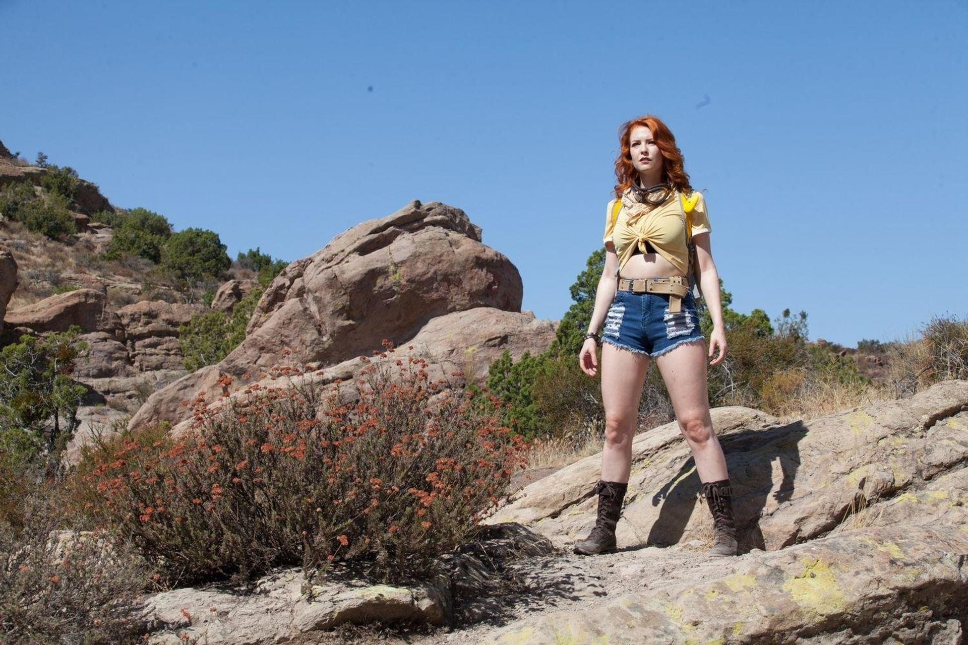 Eine Frau in Shorts und Stiefeln steht auf Felsen. Der Hintergrund zeigt Berge und blauen Himmel.