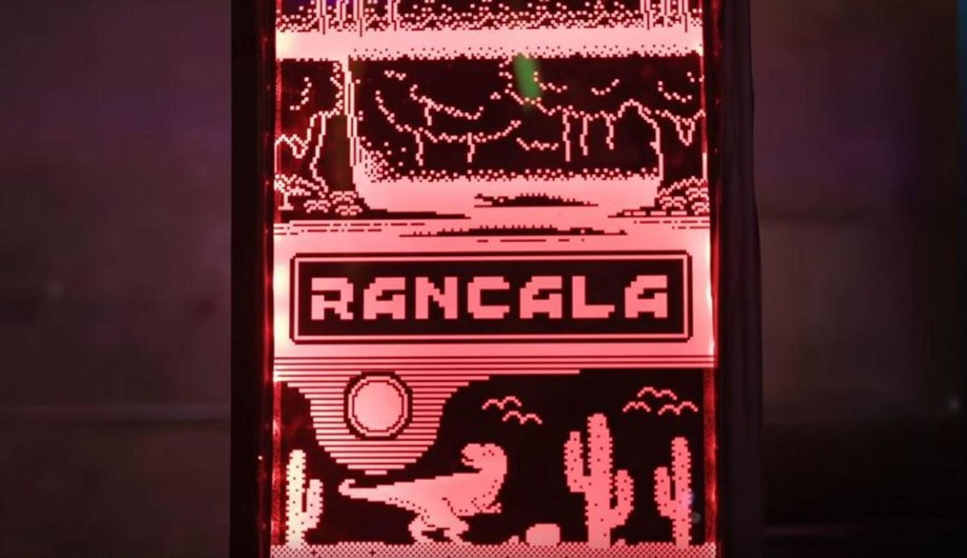 Rotes Neon-Schild mit Wüstenlandschaft und dem Namen 'Rancala'. Enthält einen Dinosaurier, Kakteen und Berge.