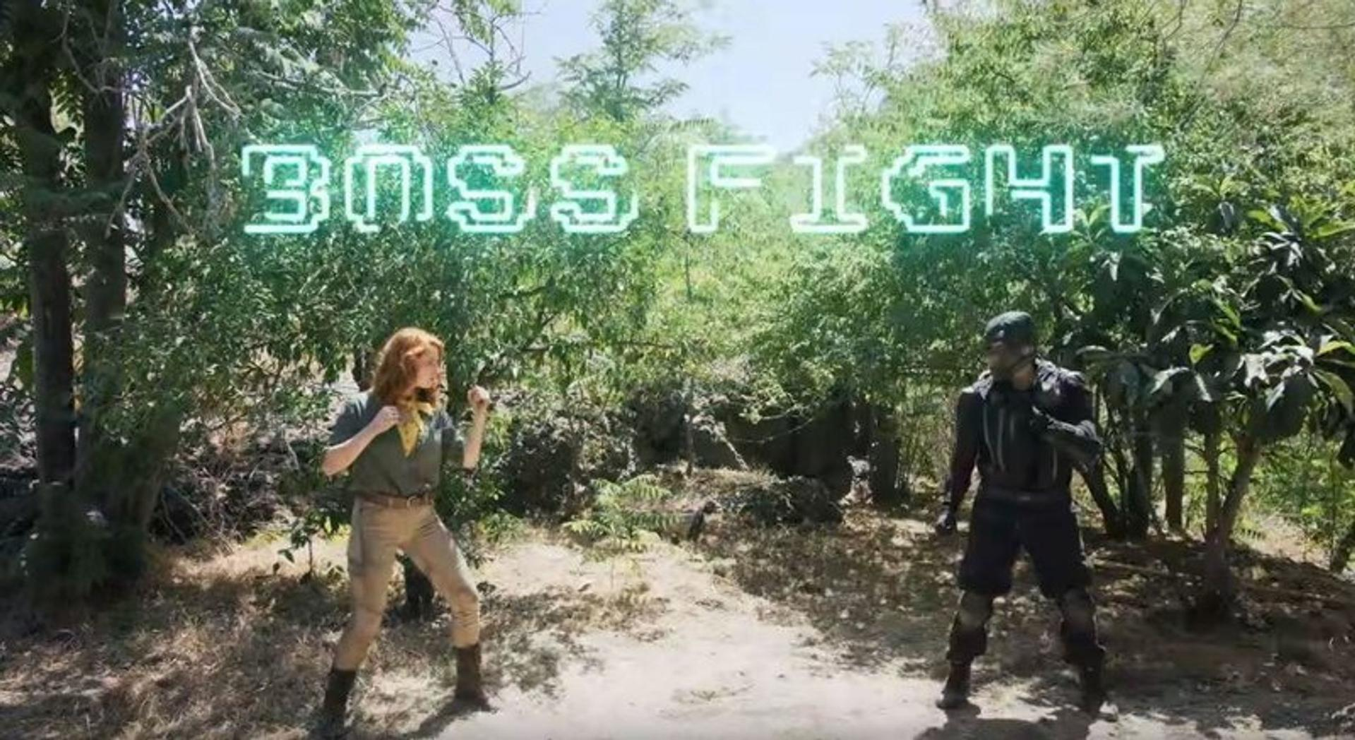 Eine Frau und ein Mann stehen sich in einer Waldumgebung gegenüber. Neon-Schild mit der Aufschrift „BOSS FIGHT“. Die Frau hat rotes Haar, der Mann trägt schwarze Ausrüstung.