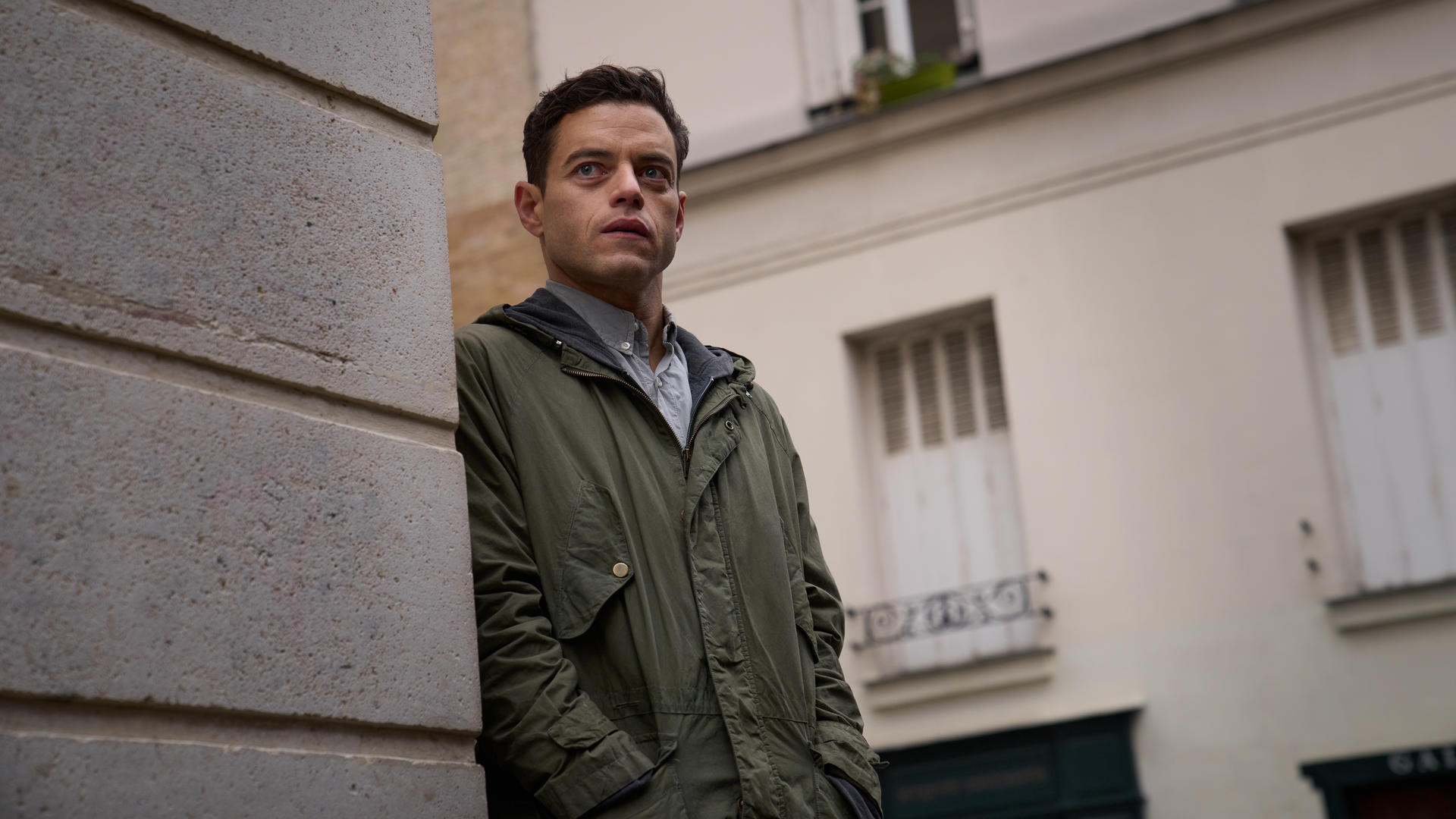 Rami Malek in einer grünen Jacke steht in der Nähe einer Steinmauer. Gebäude und Himmel im Hintergrund.