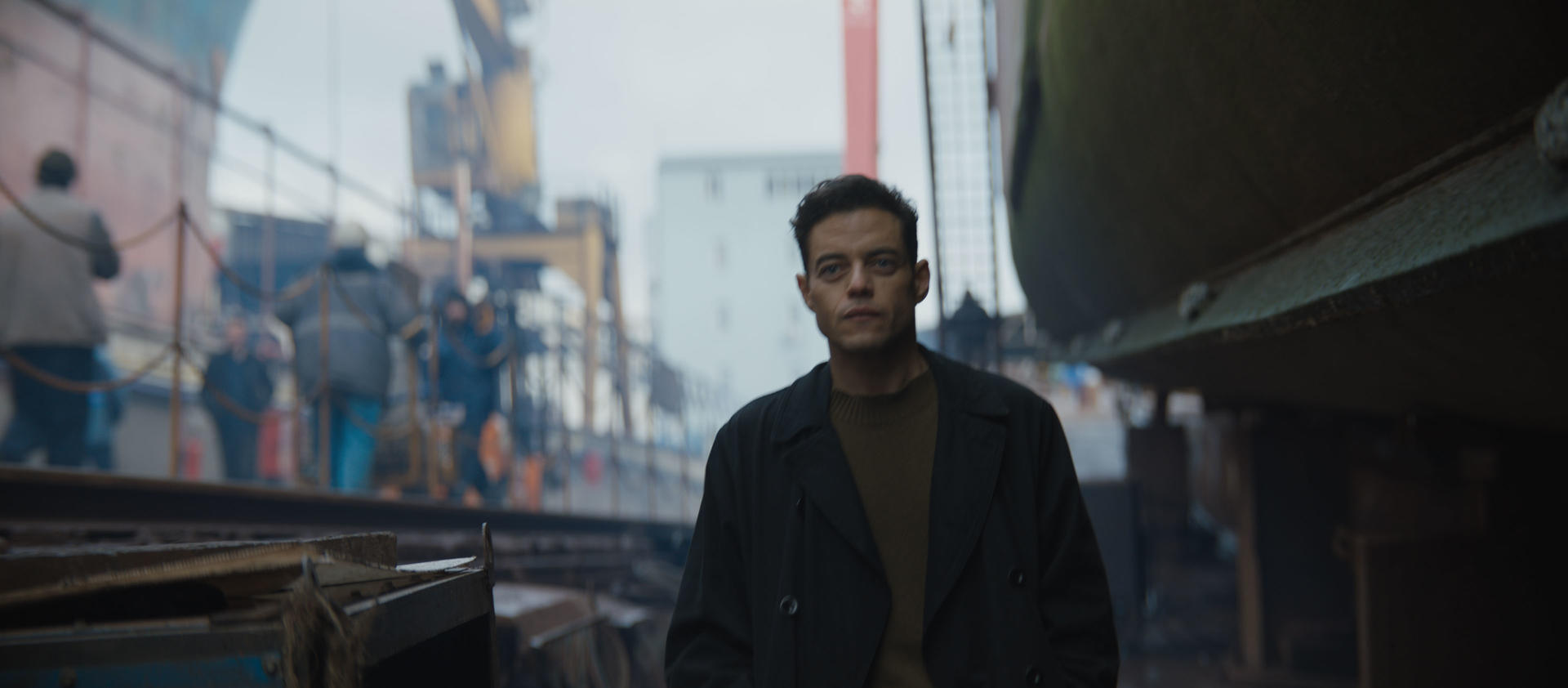 Rami Malek in einem dunklen Mantel steht in einer Werft. Arbeiter und Boote im Hintergrund.