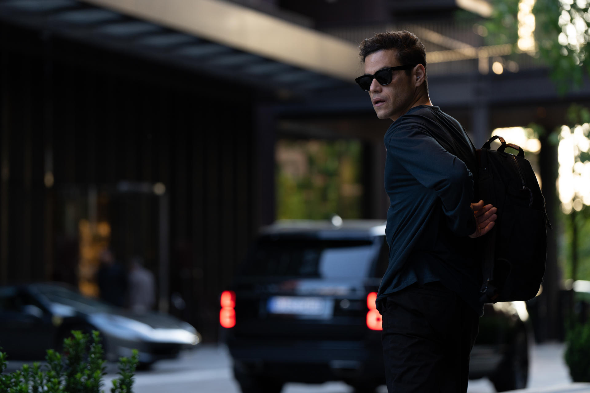 Rami Malek mit Sonnenbrille und Rucksack geht in der Nähe eines schwarzen Autos. Gebäude im Hintergrund.