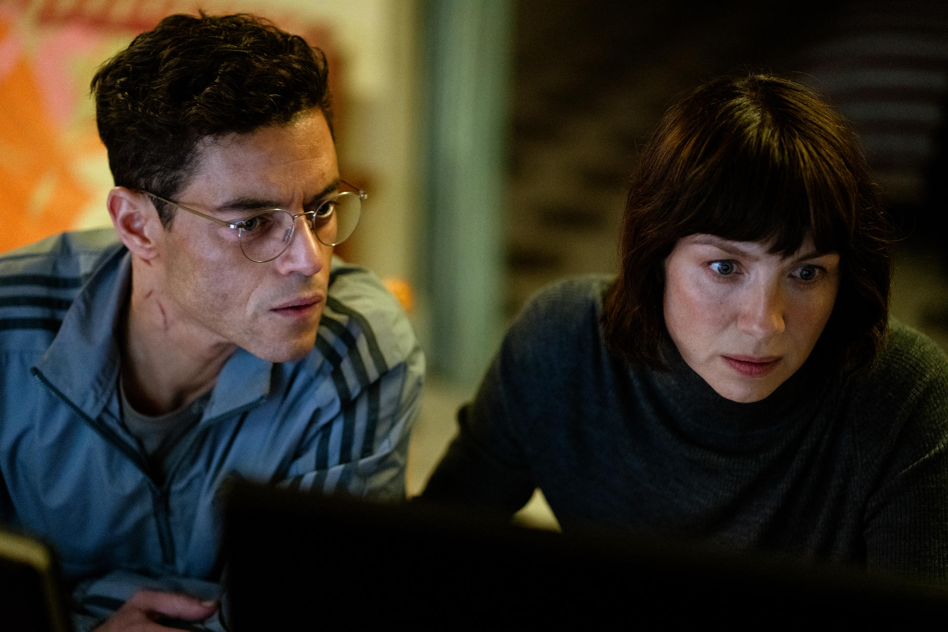 Rami Malek und eine Frau schauen auf einen Computerbildschirm. Beide sind drinnen und konzentriert.
