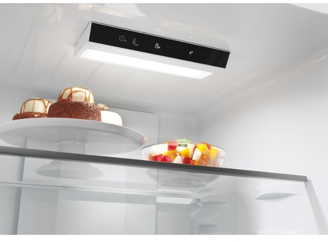 À l'intérieur d'un réfrigérateur : des gâteaux et une salade de fruits sont sur les étagères, éclairés par une lumière.