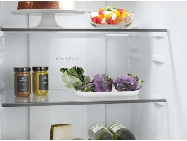 Intérieur du réfrigérateur avec aliments : bocaux, légumes, salade, gâteau. Étagères visibles.