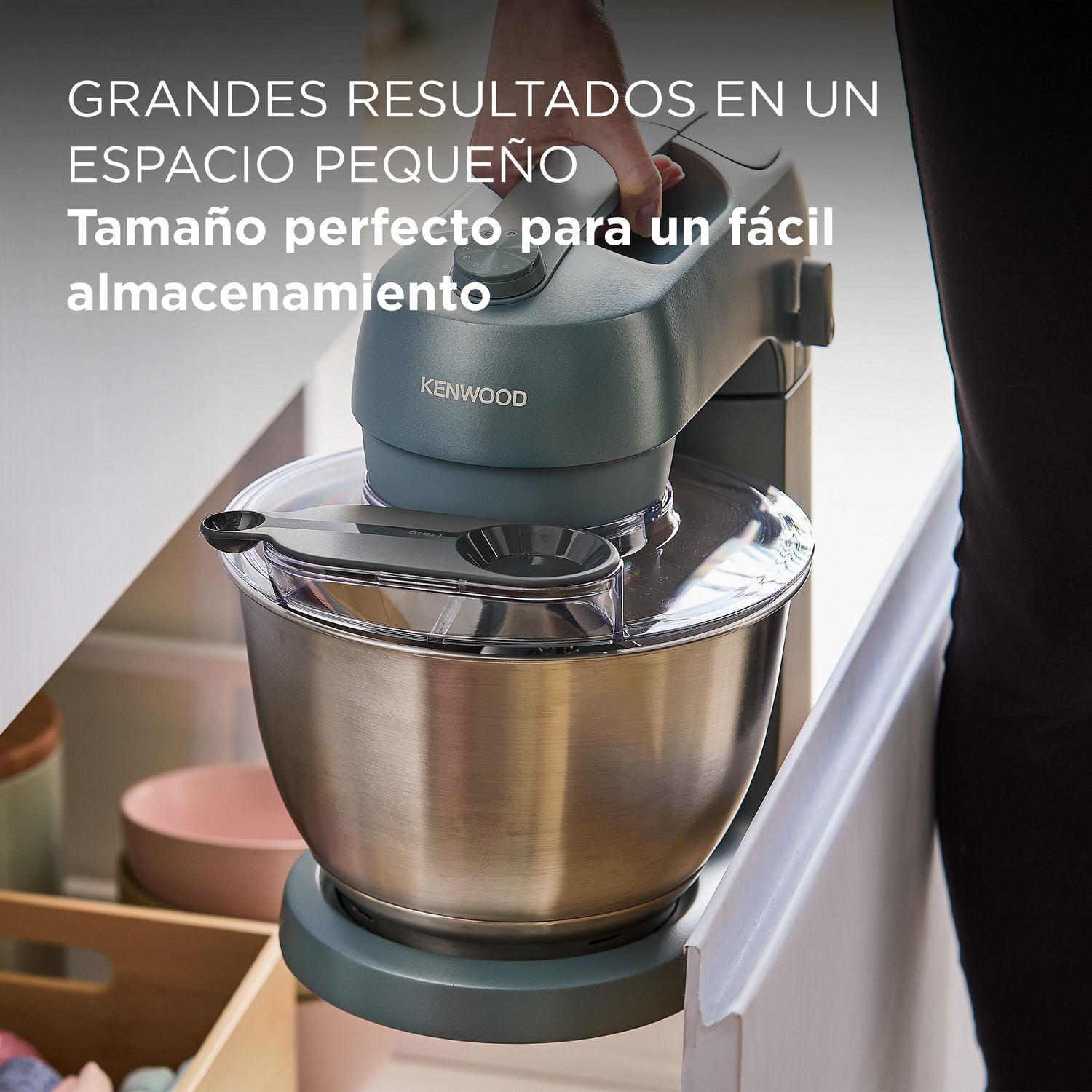 Una persona usando una batidora Kenwood azul. Está sobre una encimera. La batidora tiene un protector contra salpicaduras y una cuchara.